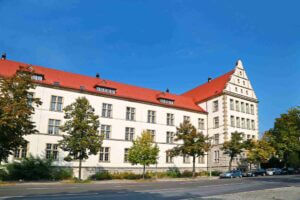 Weißes Gebäude mit rotem Dach vor blauem Himmel aņ Straße mit Bäumen