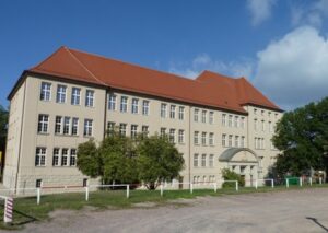 Weißes großes Schulgebäude mit rotem Dach, blauem Himmel und Hof davor